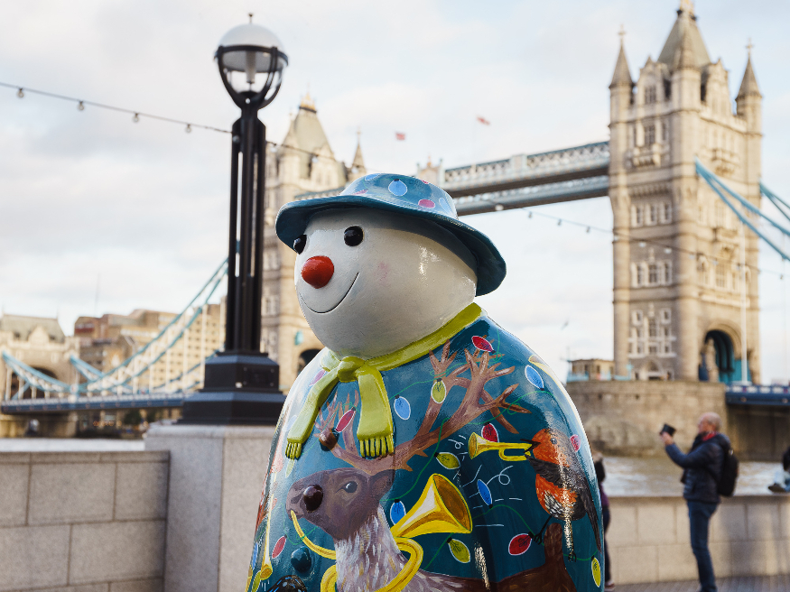 A The Snowman sculpture with a reindeer painted on its chest, in front of Tower Bridge