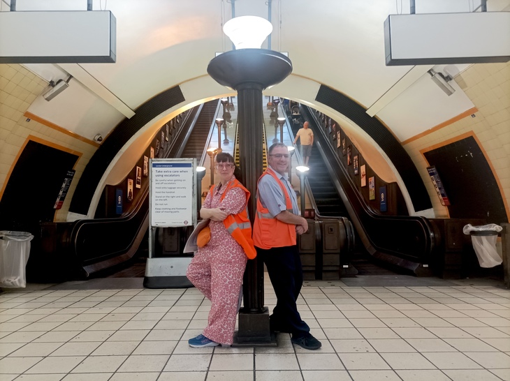 Two people in high vis posing at the bottom of an escalator