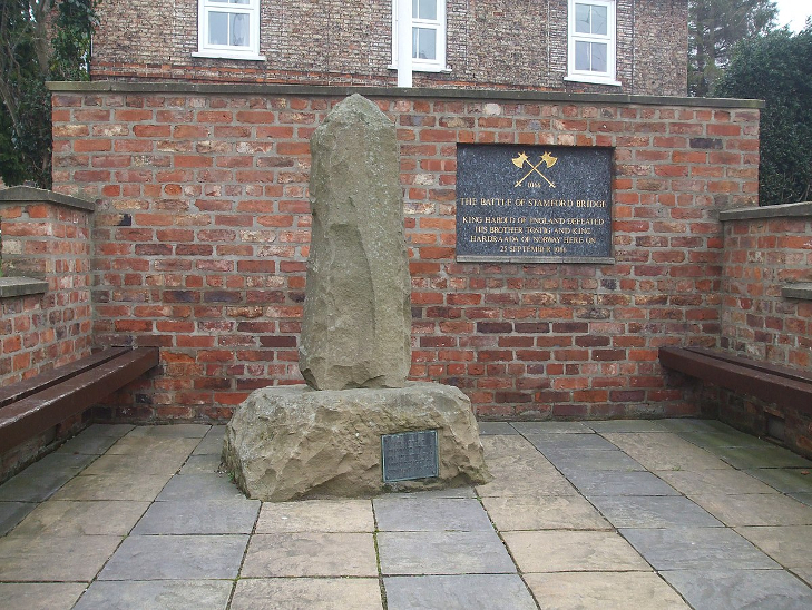 A stone monument, with a red brick wall behind it, holding a plaque in commemoration of the Battle of Stamford Bridge in 1066.