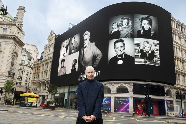 A man standing in front of the Piccadilly Circus screen, covered in black and white pics of celebs with their eyes shut