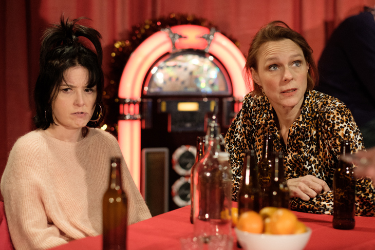 Two women sit at a table with beer bottles looking unhappy - a jukebox is in the background