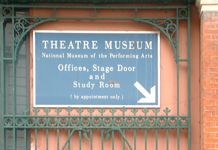 A blue sign mounted on a red wall behind a green gate. It says ' Theatre Museum National Museum of the Performing Arts Offices, Stage Door and Study Room (by appointment only)'