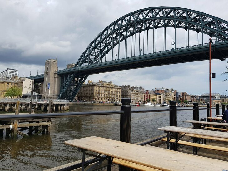 The river tyne with the magnificent Tyne Bridge can be seen. The photo is taken from the perspective of someone sitting at a table just next to the bridge on the quayside. 