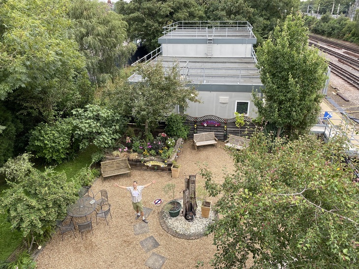 An aerial view of a small garden with gravel and shrubs. A person stands in the middle holding out their arms