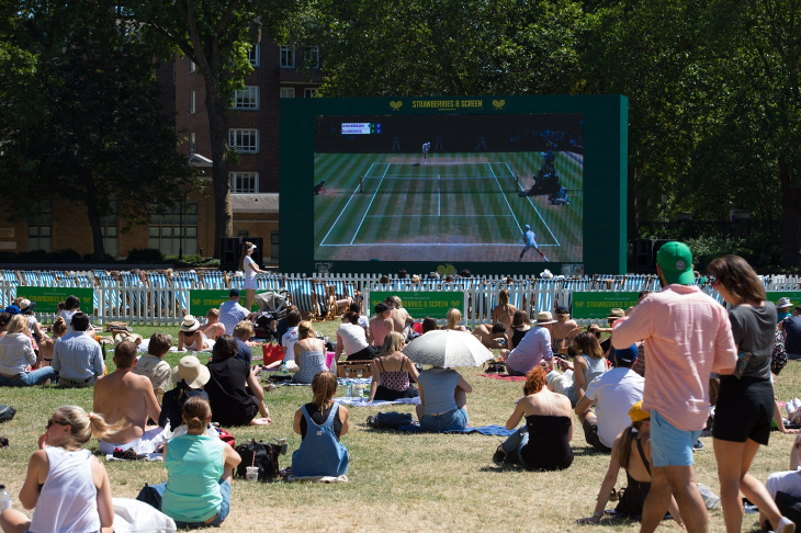 People sitting on grass in the sunshine watching tennis on a large screen