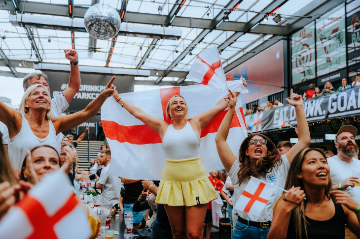 A crowd waving and wrapped in England fans watching the screen at Boxpark