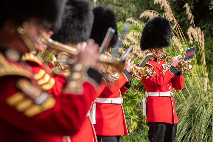 Scots Guards playing musical instruments in a garden, wearing red tunics and black bearskin hats.