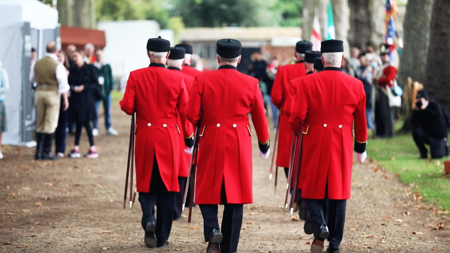 A group of Chelsea Pensioners in their red coats, walking away from the camera through the garden of the Royal Hospital Chelsea.
