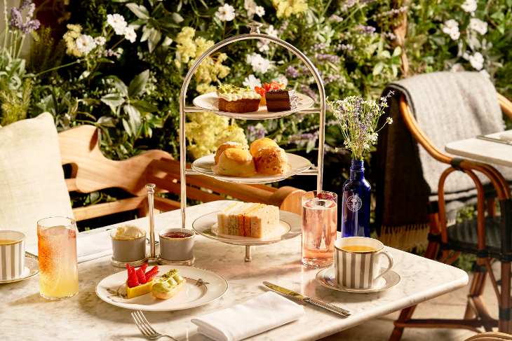 An afternoon tea stand full of food on a table, with a wall of flowers and plants in the background.