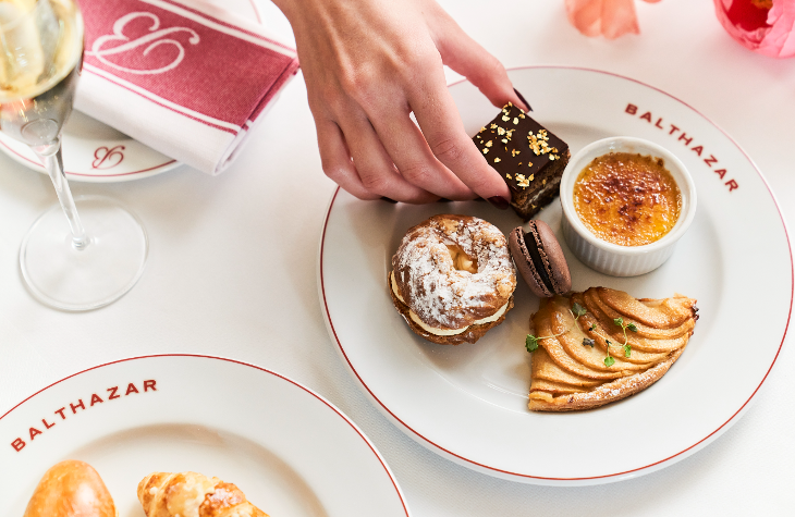 A Balthazar plate containing five desserts, including a creme brulee and a Paris Brest. A hand is reaching in to take one of the items.
