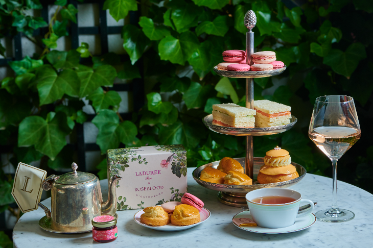 A metal three-tiered stand filled with scones, sandwiches, and pink macarons. There's also a teapot, teacup, box of macarons and glass of rose on the table.