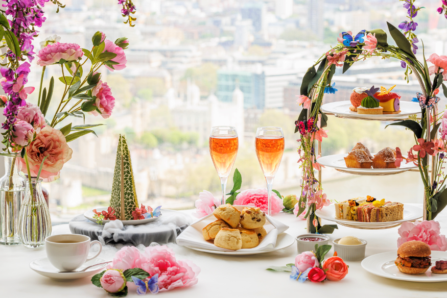 A table laid out for afternoon tea with tiered stand covered in flowers, plates of scones, glasses of wine, and a mini edible replica of The Shard.