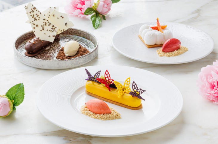 Three white plates on a marble surface, each containing a dessert. The closest one is a yellow iced cake topped with colourful butterflies. 