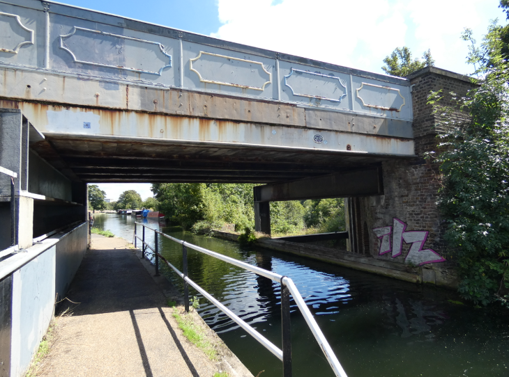 A photo taken from a canal towpath, showing the road bridge running overhead, and the railway emerging from beneath the canal.