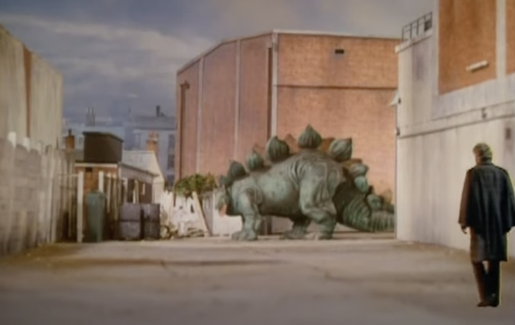 A stegosaurus wanders around some light industrial buildings while third doctor Jon Pertwee looks on 