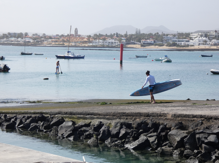 Corralejo in Fuerteventura: someone walking into the sea with a surfboard, with others on paddleboard in the background at Corralejo Harbour