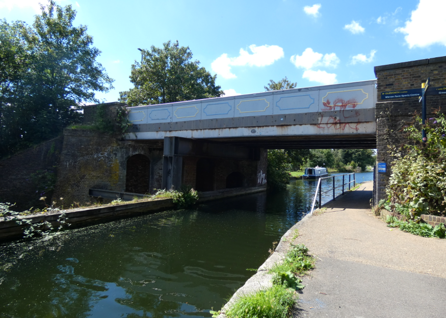 Photo taken from the towpath on the side of the canal, with a metal road bridge crossing over the top of the canal.