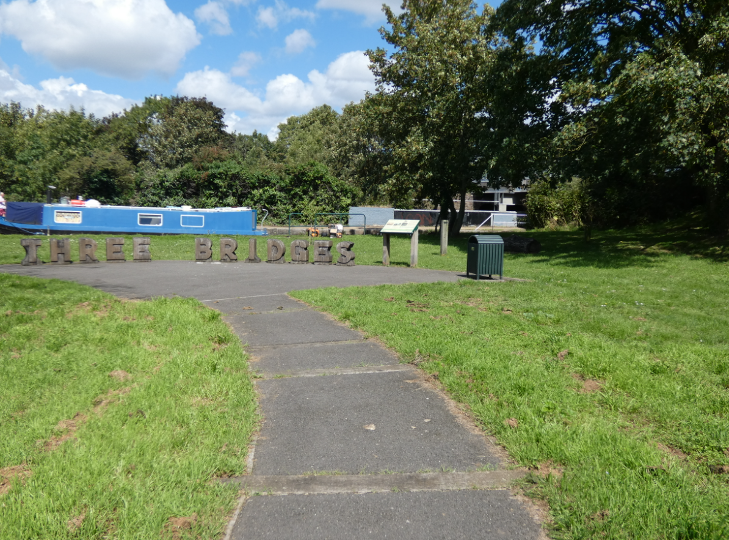 A tarmac path intersperesed with wooden railway sleepers, leading to an open area with wooden 3d letters saying 'Three Bridges'. There's a blue canal boat in the background, and the road bridge can just be seem through trees to the right.
