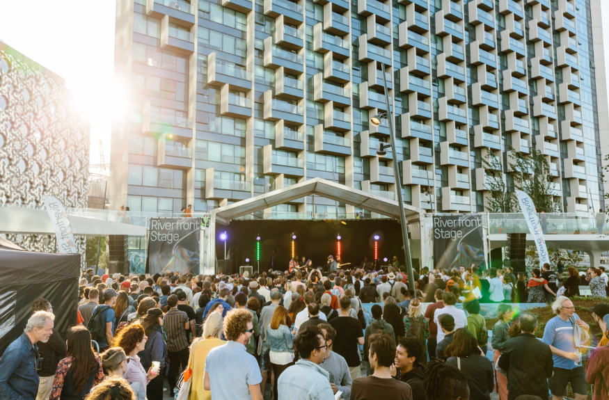 Crowds watching acts on an open-air stage in front of a tall building at Turning Tides festival at Greenwich Peninsula