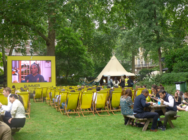 A big screen against a backdrop of trees, with people sitting on wooden picnic benches and yellow deckchairs