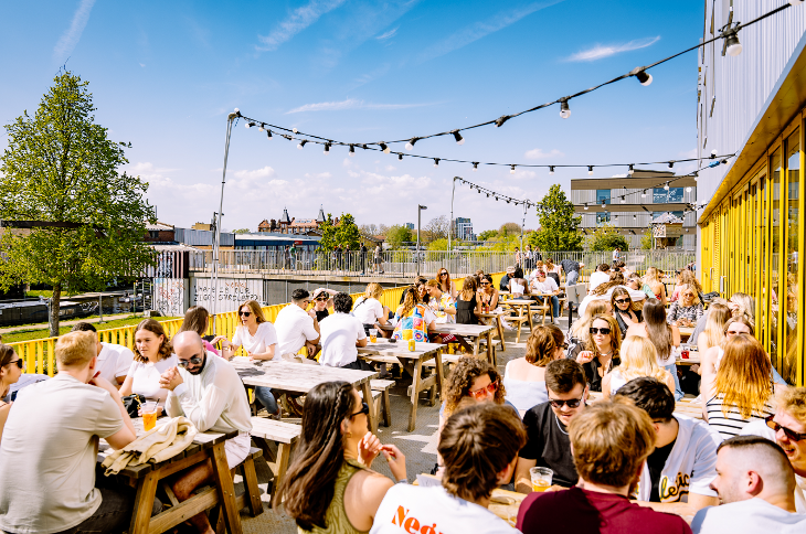 An outdoor terrace full of picnic tables with people drinking in the sunshine