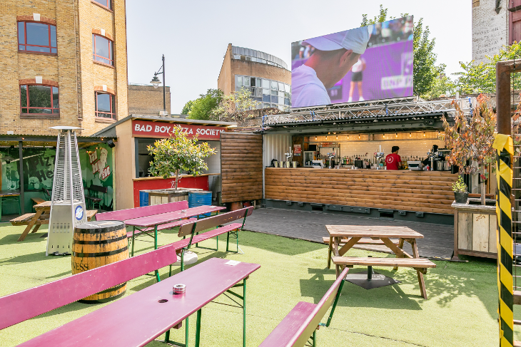 A courtyard area with picnic tables and fake grass, featuring a bar with a giant screen on its roof